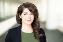 Anne, a woman with brown eyes, medium length brown hair and bangs, in a green top and black blazer, smiles with the USPTO headquarters hallway behind her.