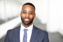 Darren, a man with short black hair, black beard, a navy suit, light blue tie, and white collared shirt, smiles in USPTO headquarters hallway.