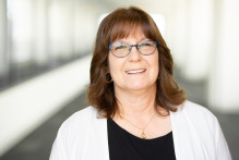 Debra, a woman with glasses, medium brown hair, gold hoop earrings and necklace, white and black shirt, smiles in USPTO headquarters hallway.