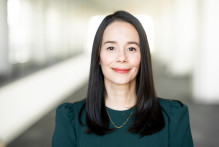 Glo, a woman with brown eyes, medium length dark brown-black hair, in an emerald green top with ruched sleeves and a gold necklace, smiles with the USPTO headquarters hallway behind her.