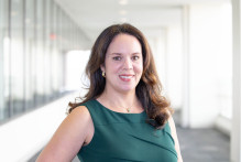  Norca, a woman with brown eyes, medium length dark brown hair, a sleeveless emerald green professional top, and small gold hoop earrings and a gold necklace with a green gem in it, smiles with the USPTO headquarters hallway behind her.