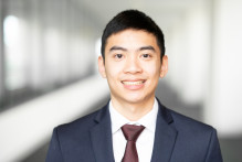 Steven, a man with black hair, in a navy blue suit, red tie, and white collared shirt, smiles with the USPTO headquarters hallway behind him.