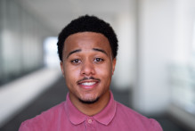 Andrew, a young man with short dark hair, wears a light red collared polo shirt while smiling in a hallway at USPTO headquarters.