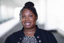 Angelica, a woman with braided hair in a bun, wears a black and white pattern blouse and dark jacket while smiling in a hallway at USPTO headquarters