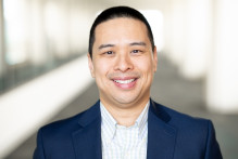 Man with short hair wearing a dark blazer and white collared work shirt smiles in a bright hallway.