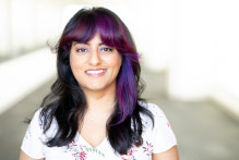 USPTO trademark examining attorney Komal V. poses in a light-filled corridor at the Alexandria, Va., headquarters. Komal wears a flowered shirt and has purple streaks in her black hair.