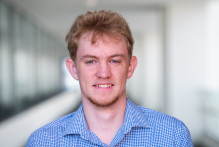 Max, a young man with short length light hair, wears a long sleeve blue and white collared shirt while smiling in a hallway at USPTO headquarters. 