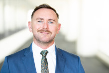 USPTO headshot of Jordan L. He wears a blue suit, white shirt, and flowered tie.