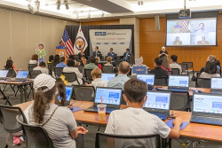 A mix of kids and adults sit in lecture style room with laptops on their desk while a woman speaks at a podium