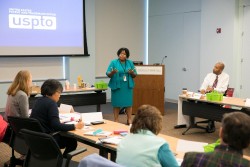 A woman standing in front of podium and speaking to men and women seated at desks.