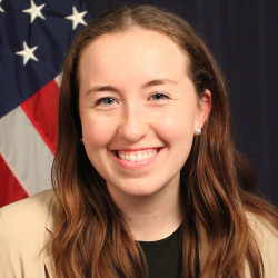 Photograph of a woman in front of an American flag smiling who has light skin, long dark hair, and is wearing a beige jacket, black shirt, and pearl earrings.