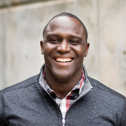 Photograph of a man smiling who has dark skin, short hair, and is wearing a grey sweater with a red and grey plaid collared shirt underneath.